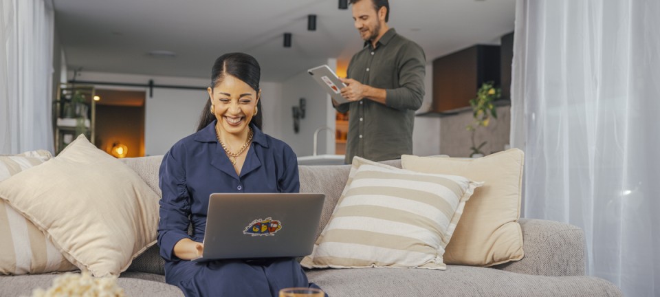 Woman smiling at her laptop on the couch while a man stands behind her
